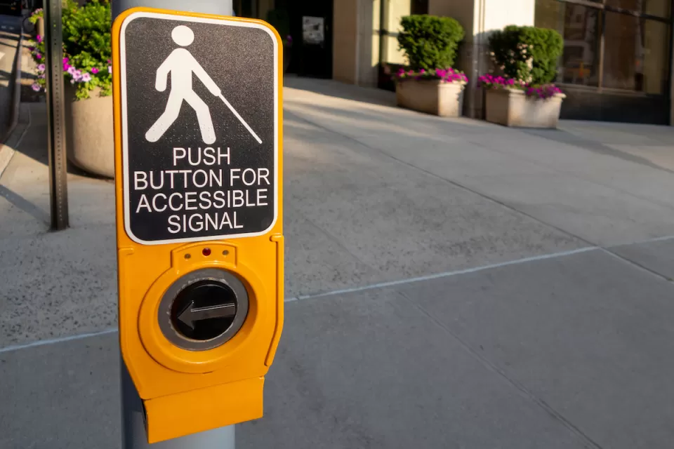 Close-up of yellow pedestrian crossing signal button with text "Push button for accessible signal" and diagram of person holding cane, indicating blindness.